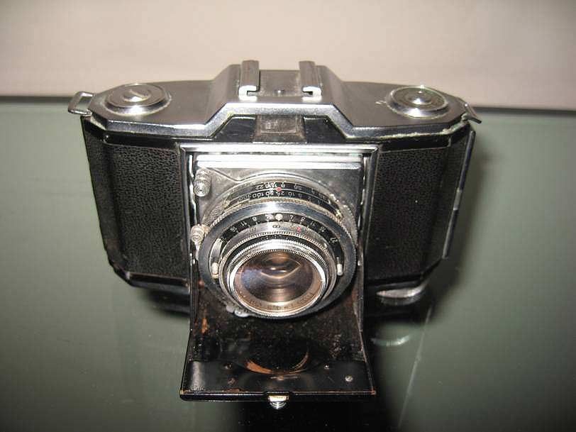 Un appareil photo argentique pliant noir vintage est posé sur une surface réfléchissante, photographié en plongée.