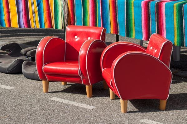 Deux petites chaises rouge vif de style vintage, avec des passepoils blancs, sont posées sur l'asphalte devant un fond en tissu rayé coloré.
