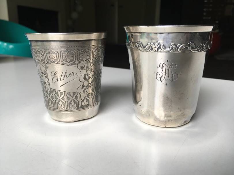 Deux coupes en argent finement ouvragées posées sur une surface blanche, l'une gravée du nom « Esther » et l'autre d'un monogramme.