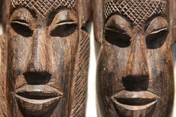 Deux masques en bois sombre sculpté, aux fronts texturés et aux traits stylisés, sont placés côte à côte.