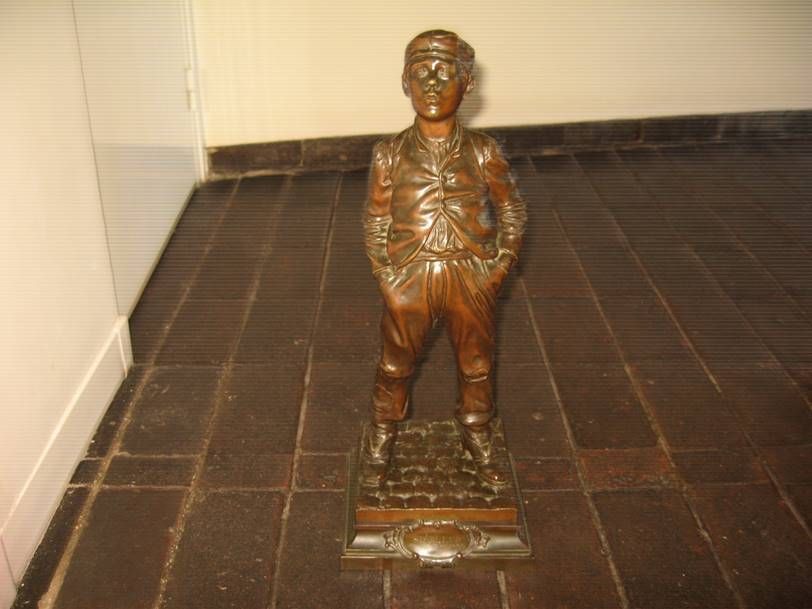 Une sculpture aux tons bronze représentant un personnage debout, les mains dans les poches, sur un socle carré, elle-même posée sur un sol carrelé sombre.