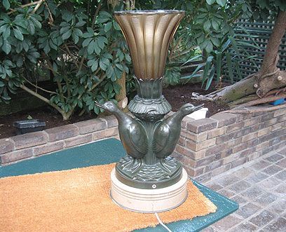 Une lampe vintage en bronze avec un sommet évasé et un pied orné de trois canards stylisés sur un support circulaire à plusieurs niveaux.