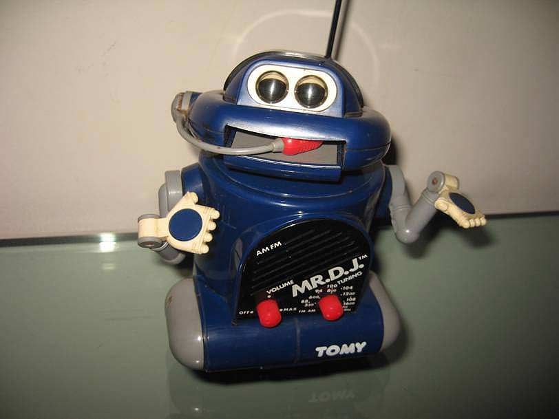 Un jouet robot vintage bleu Tomy Mr. DJ avec un casque, une antenne et deux boutons rouges, posé sur une surface réfléchissante.