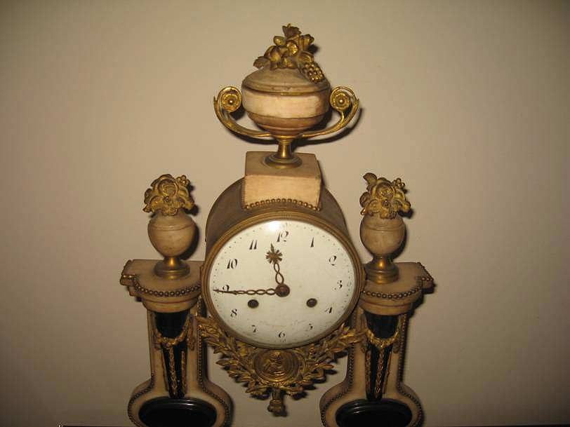 Une horloge ancienne et ornée, dotée d'un cadran blanc à chiffres noirs, de colonnes en marbre et de délicates décorations dorées.