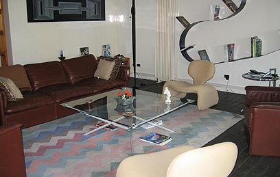 Un salon avec un canapé en cuir marron, des fauteuils beiges, une table basse en verre et un tapis à motifs sur un sol foncé.