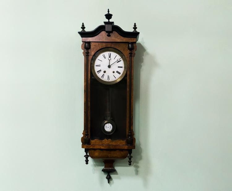 Une horloge murale en bois foncé avec un cadran blanc circulaire, des chiffres romains et un pendule suspendu, sur un mur vert pâle.