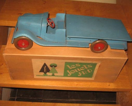 Un camion jouet bleu à plateau et à roues rouges repose sur une boîte en bois étiquetée les Jouets JRD avec un logo représentant un arbre.