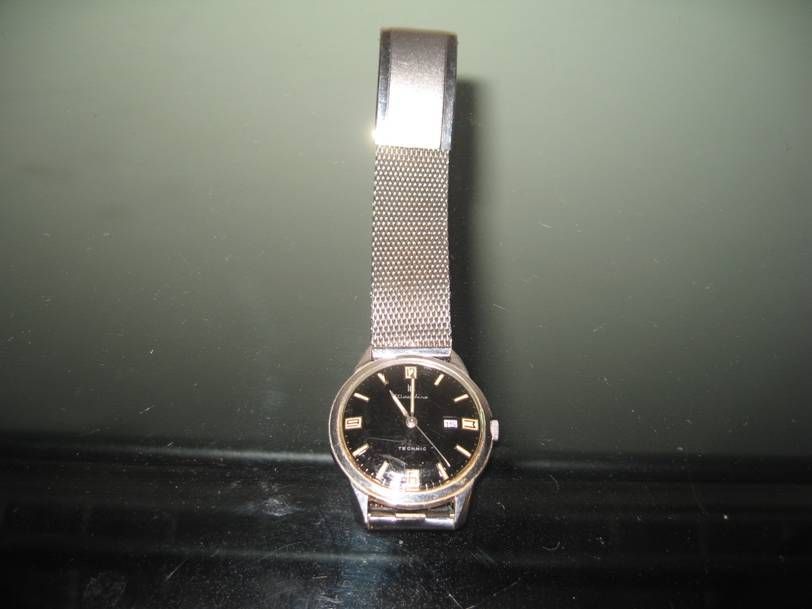 Une montre-bracelet à cadran noir et bracelet en maille métallique argentée posée sur une surface sombre.