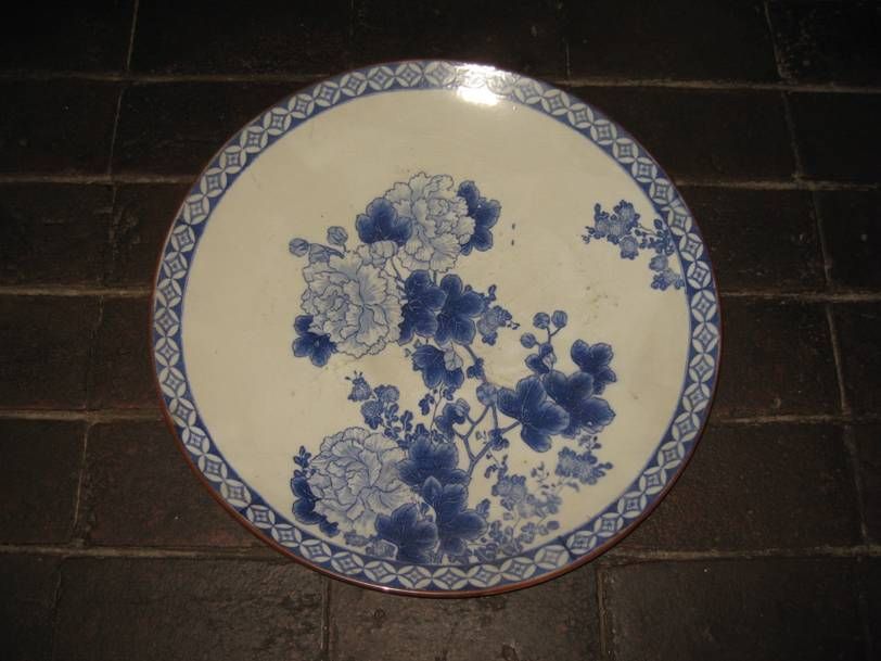 Une assiette ronde en céramique blanche ornée de motifs floraux bleus et d'une bordure géométrique, posée sur une surface carrelée foncée.