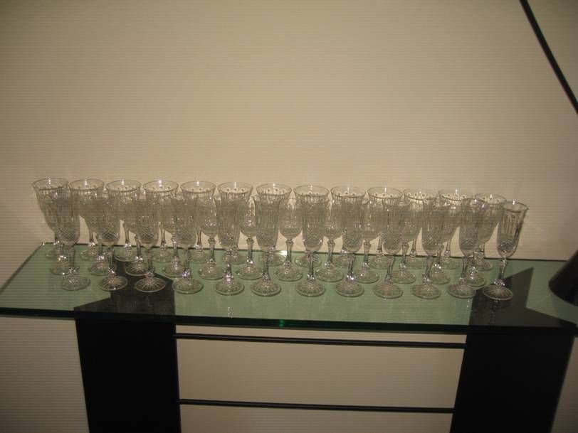 Une longue rangée de verres en cristal transparents à pied, disposés sur une console en verre contre un mur uni.