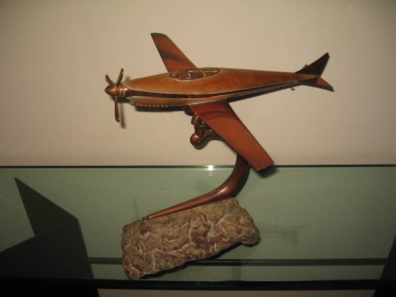 Un modèle réduit d'avion couleur bronze avec une hélice, monté sur un support métallique incurvé lui-même posé sur un socle texturé imitant la pierre.