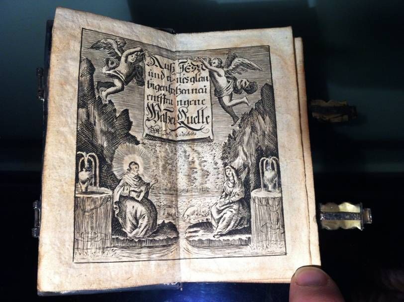 Un livre ancien ouvert laisse apparaître une gravure représentant des chérubins, des figures religieuses et un texte calligraphique central.