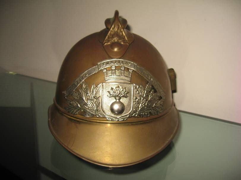 Un casque de pompier français ancien en laiton, orné d'un emblème représentant une grenade enflammée sur une surface en verre.