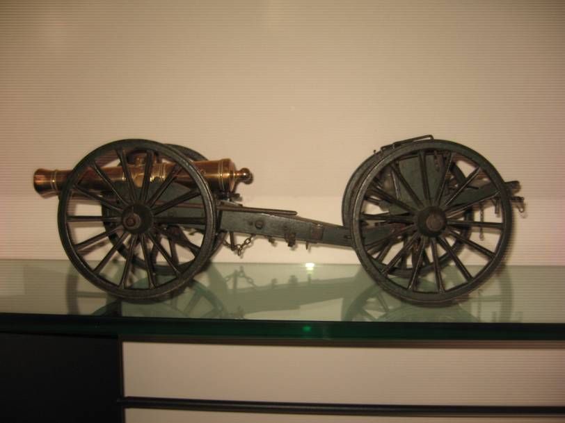 Un canon miniature en laiton à roues noires, posé sur une étagère en verre sur fond blanc.