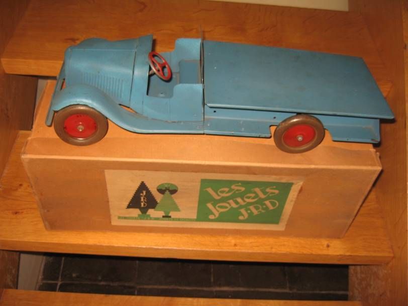 Un camion jouet vintage bleu à roues rouges, posé sur sa boîte en bois d'origine étiquetée « les JOUETS JRD », sur un escalier.