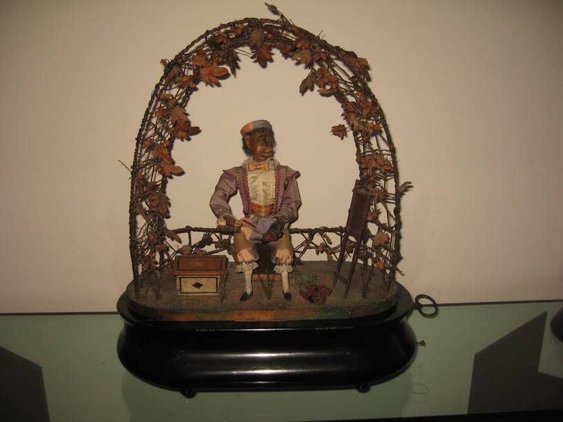 Un automate ancien représentant un personnage peignant sur un chevalet sous une arche en fil de fer ornée de feuilles d'automne séchées.