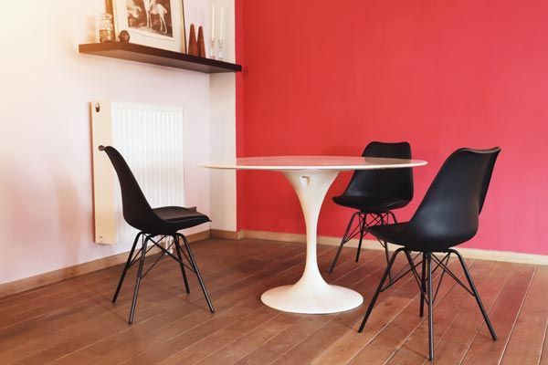 Une table blanche de style tulipe avec trois chaises noires sur un parquet, devant un mur blanc et un mur rouge vif.