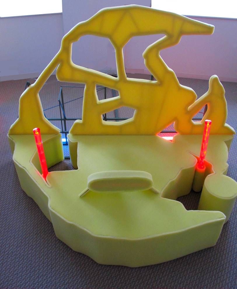 Une structure abstraite jaune vif, semblable à de la mousse, avec des découpes et deux lumières verticales rouges incandescentes, sur un sol recouvert de moquette.