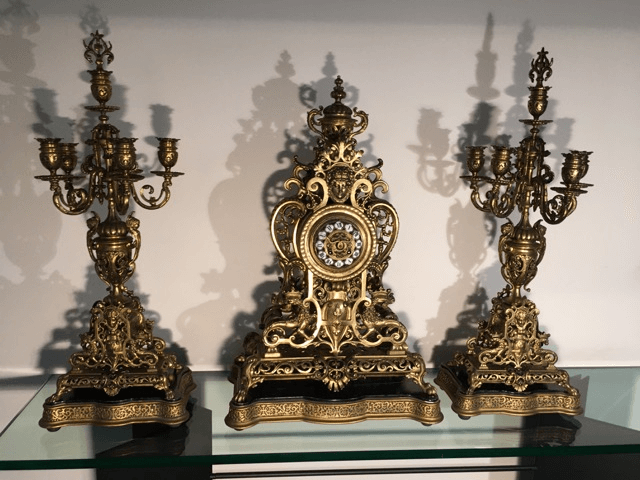 Une pendule de cheminée ancienne et ornée, accompagnée de deux candélabres à cinq bras assortis en métal doré, posée au centre d'une étagère en verre.