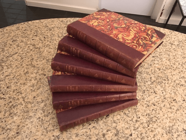 Une pile de six livres anciens, à la reliure en cuir bordeaux foncé et à la couverture à motifs marbrés, posés sur une surface mouchetée.