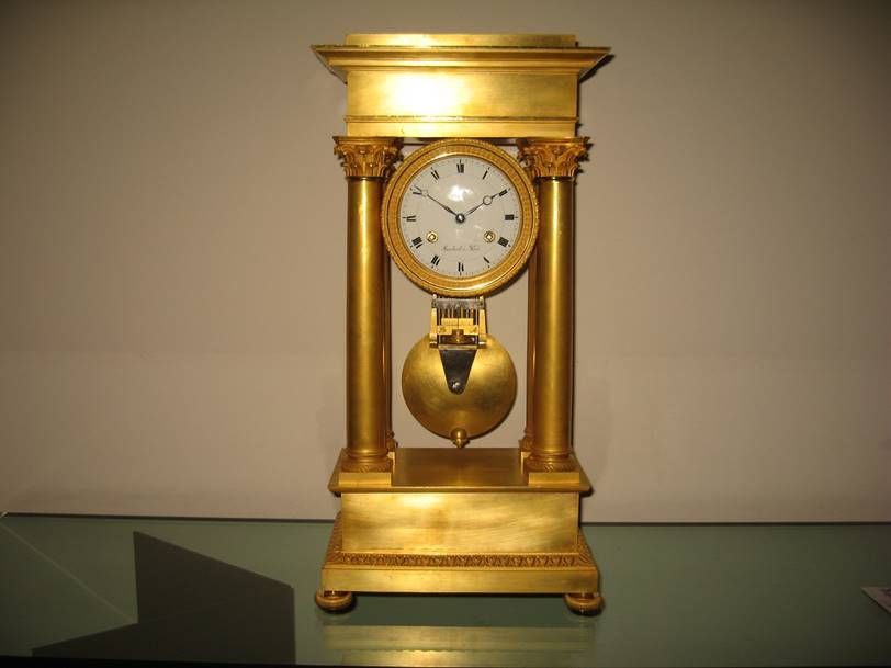 Une grande pendule de cheminée en laiton doré, à cadran blanc, deux colonnes et pendule visible, posée sur une table.