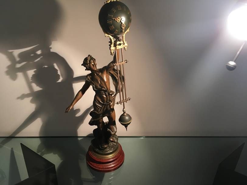 Une statue en bronze soutient une horloge circulaire, projetant une longue ombre sur un mur nu.