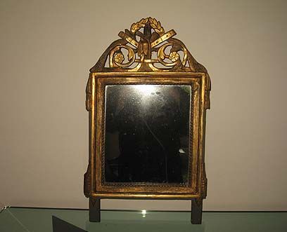 Un petit miroir rectangulaire orné, de couleur dorée, avec un blason sculpté élaboré sur le dessus, monté sur deux pieds verticaux sombres.