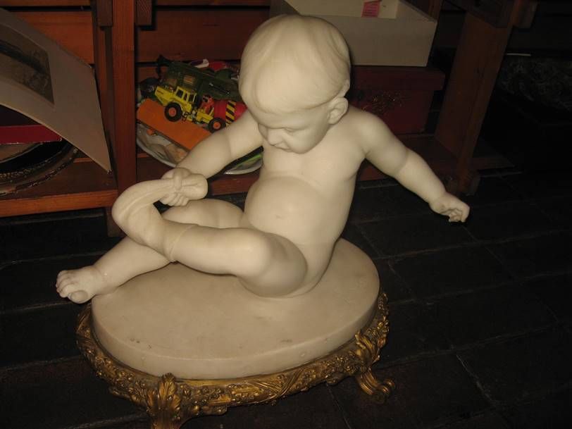 Une statue en marbre représentant un enfant assis sur un socle doré orné, tenant son pied, posée sur une étagère en bois.