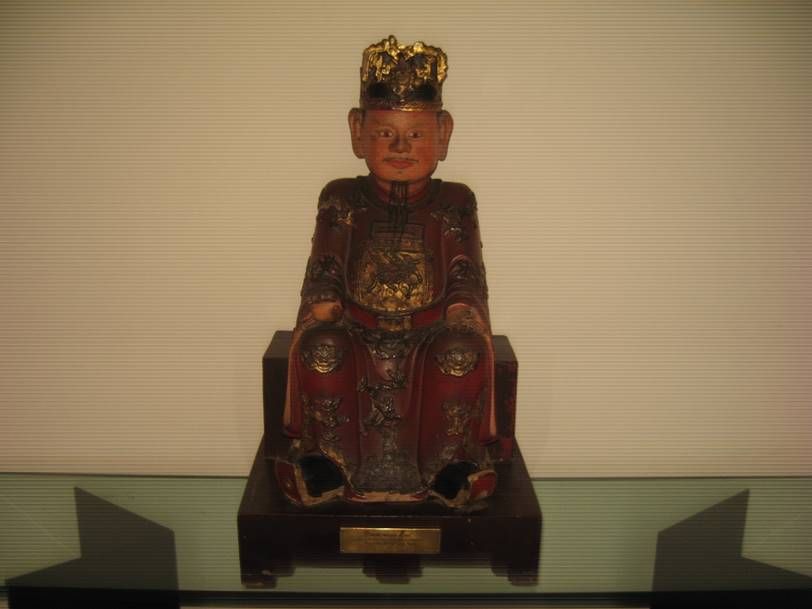 Une figure assise, vêtue d'une robe rouge et coiffée d'une couronne dorée, placée sur un socle en bois sombre devant un mur neutre.