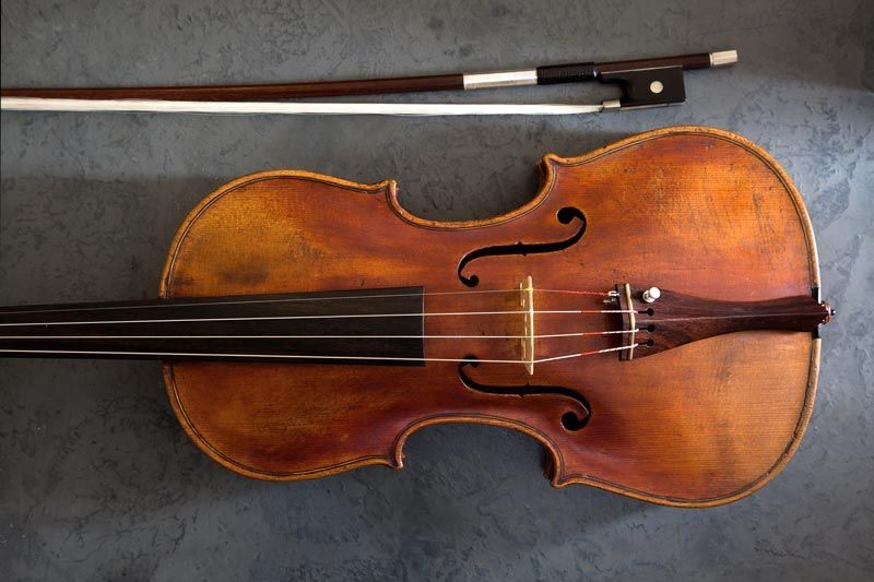 Un violoncelle en bois, son archet posé au-dessus, sur un fond gris foncé texturé.