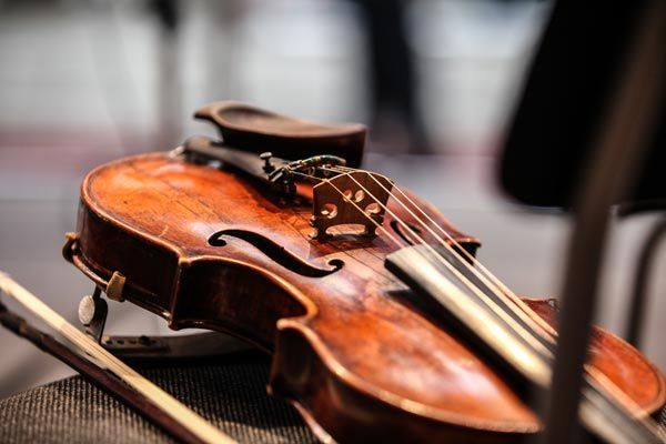 Un violon en bois brun usé repose sur une surface en tissu à côté de son archet, avec un orchestre flou en arrière-plan.