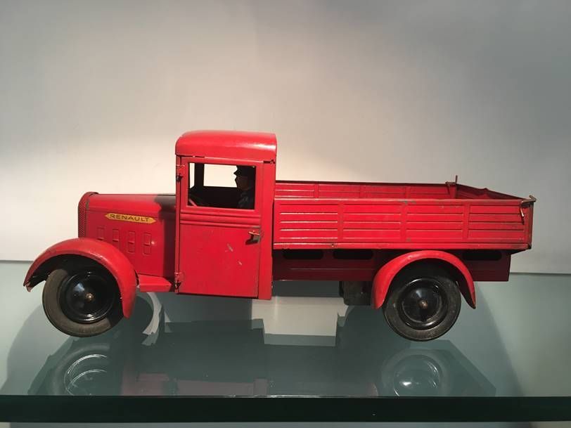 Un camion miniature vintage en métal rouge est posé sur une surface vitrée réfléchissante, sur un fond clair uni.