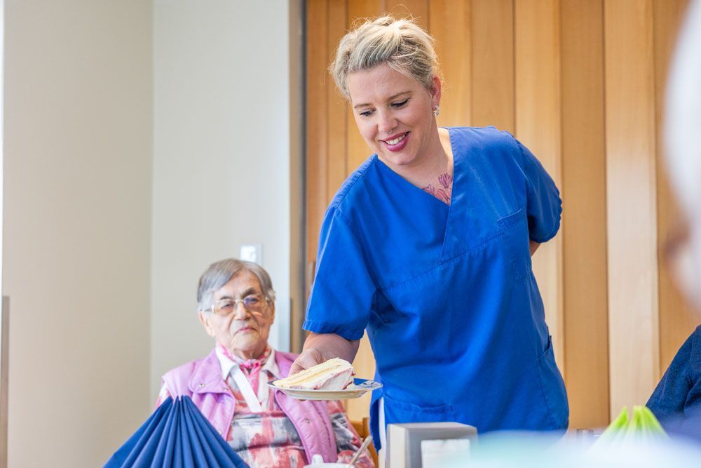 Eine Krankenschwester serviert einer älteren Frau, die an einem Tisch sitzt, Essen.