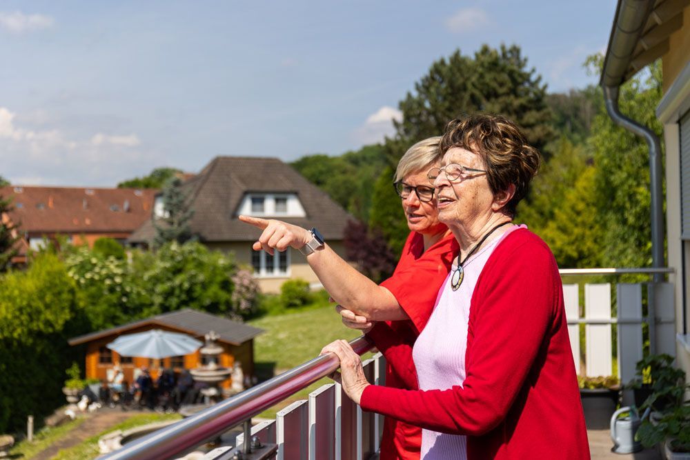Zwei ältere Frauen stehen auf einem Balkon und blicken auf einen Garten.