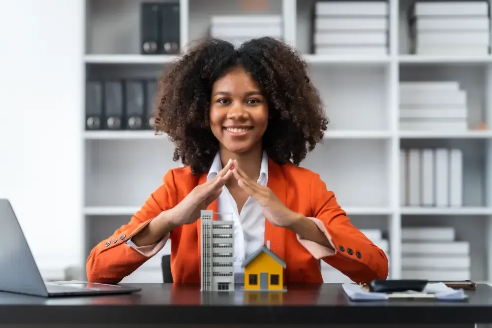 Uma mulher negra sorridente usando um blazer laranja protege prédios em miniatura com as mãos em um escritório.