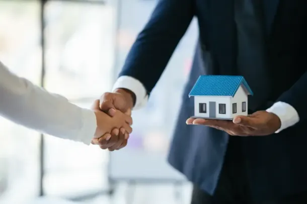 Uma pessoa de terno aperta a mão de outra, segurando uma casa em miniatura, provavelmente um negócio imobiliário.