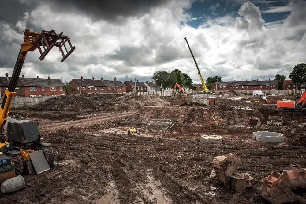Canteiro de obras com maquinário pesado e céu nublado; sujeira e detritos cobrem o chão, casas residenciais ao fundo.