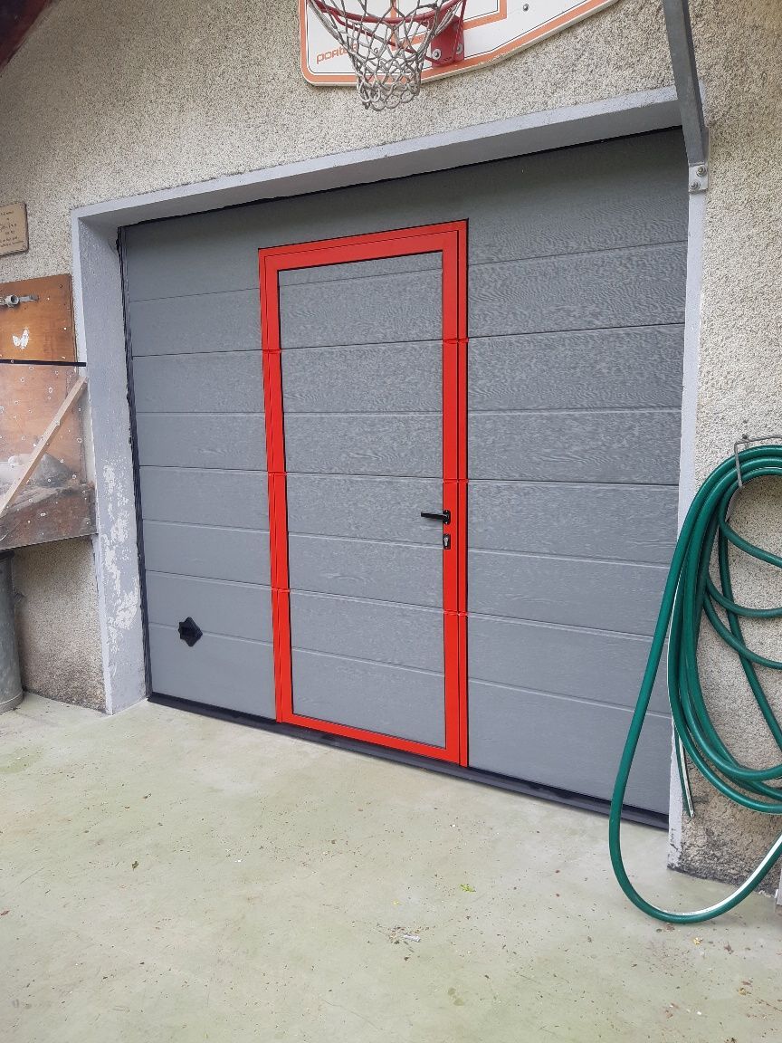 Porte de garage grise avec une porte piétonne à contour rouge, à côté d'un tuyau d'arrosage vert enroulé.