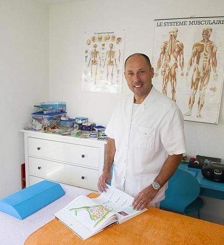 Un homme en blouse blanche sourit, debout dans une pièce remplie de planches d'anatomie, un livre ouvert à la main