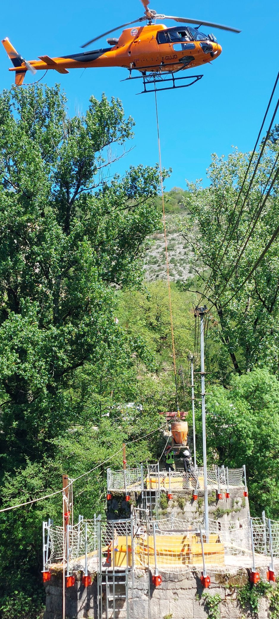 Un hélicoptère orange soulève du matériel depuis un échafaudage dans une forêt luxuriante et verte.