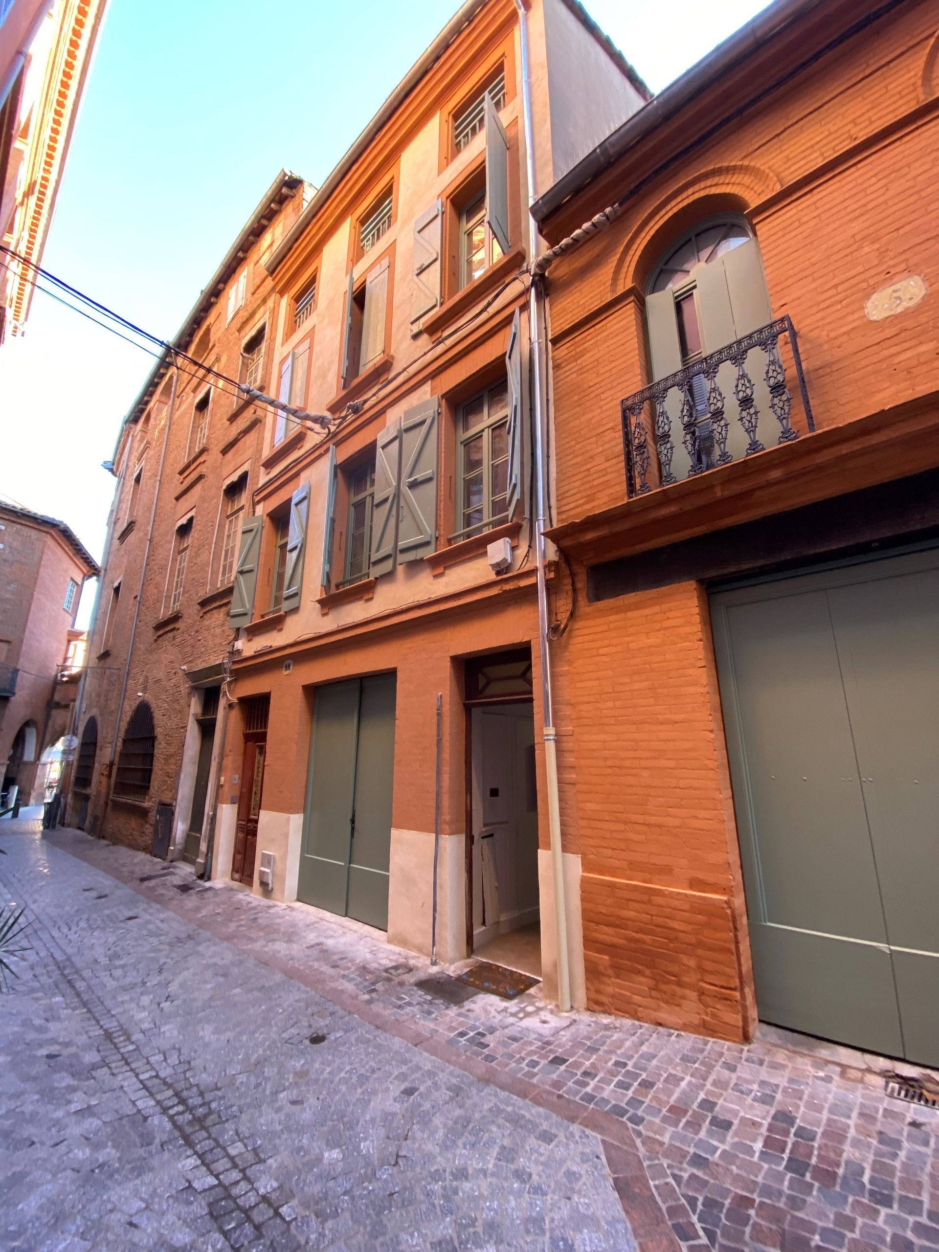 Des bâtiments en briques rouges bordent une étroite rue pavée. Quelques-uns ont des portes et des volets verts.