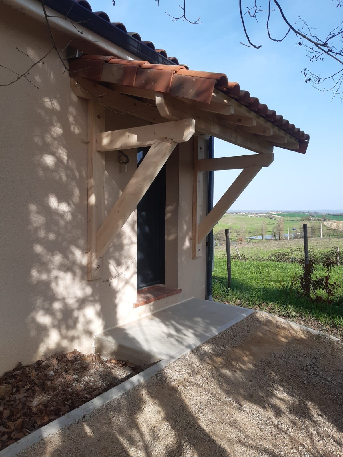 Auvent en bois au-dessus d'une porte; bâtiment beige, toit en tuiles rouges, chemin de gravier et champ vert.