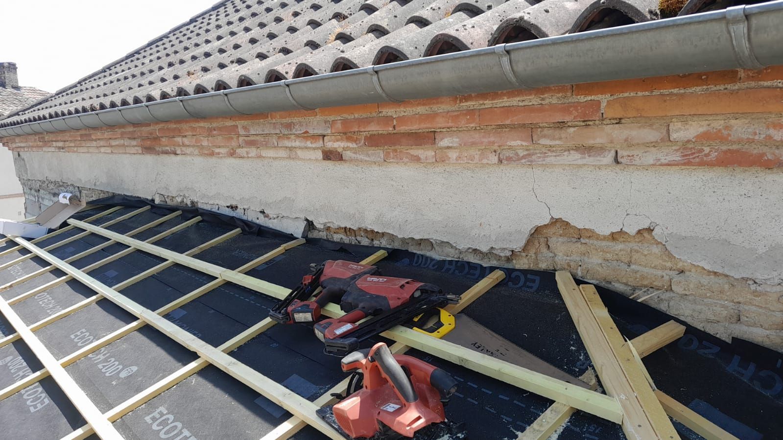 Travaux de toiture en cours : Outillage sur sous-couche noire avec supports bois près de toiture en briques et tuiles.
