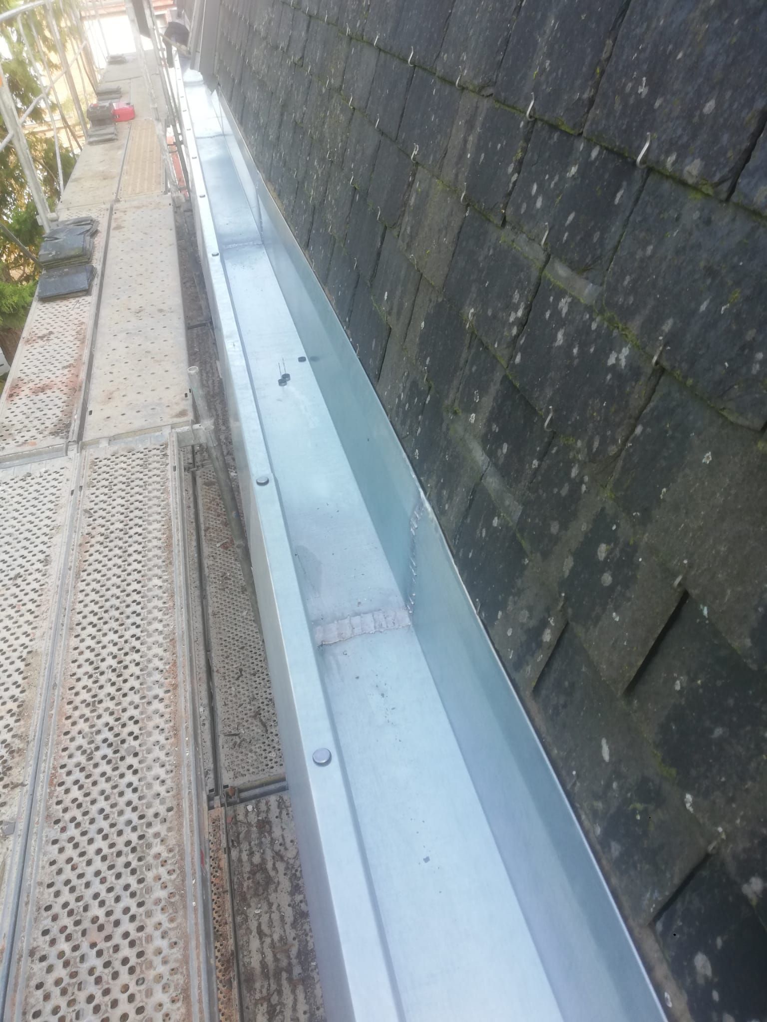 Nouvelle gouttière en métal brillant installée sur un toit en tuiles foncées à côté d'un échafaudage.