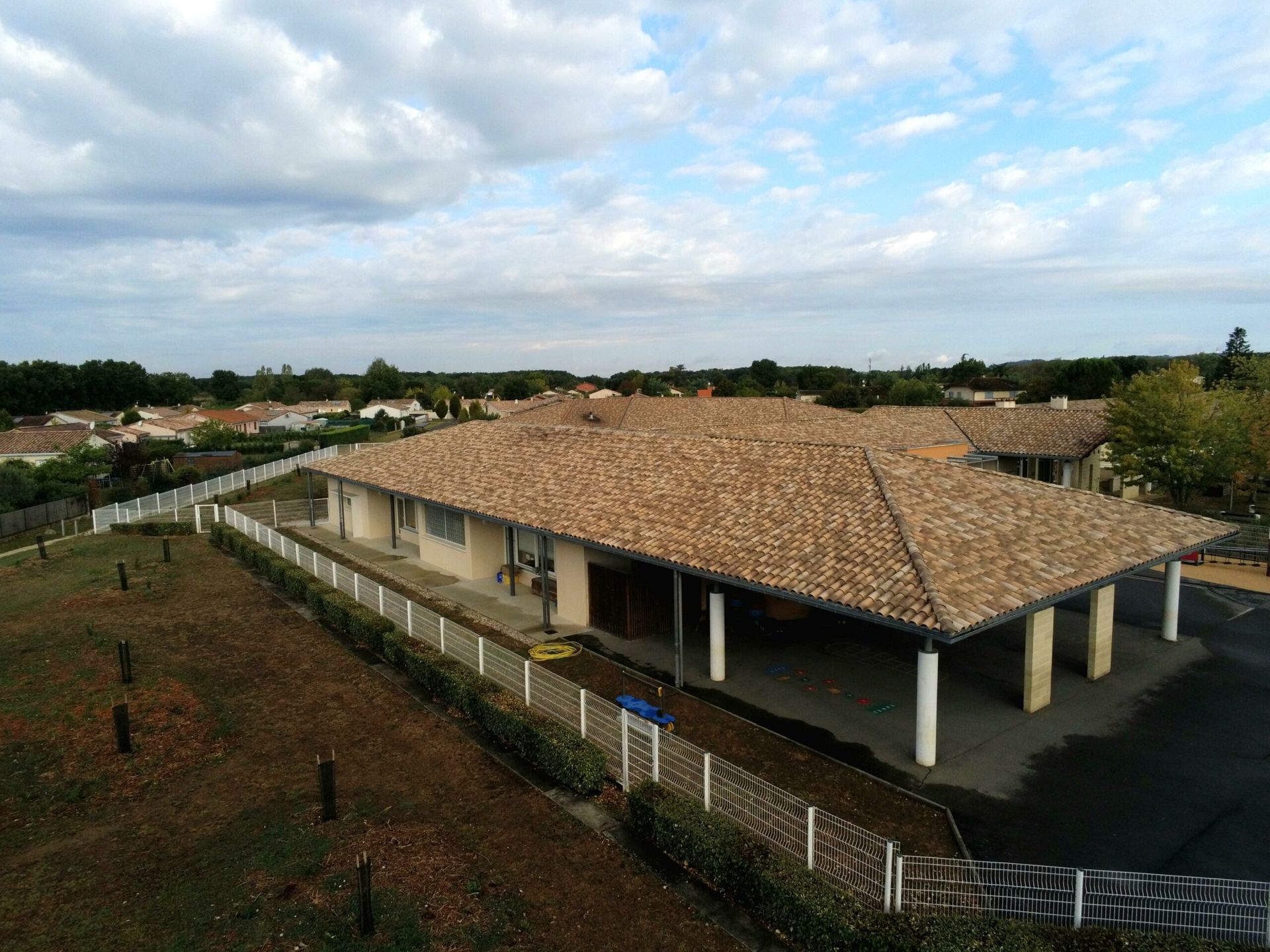 Bâtiment avec un toit en tuiles brunes et une entrée couverte à colonnes, situé dans une zone herbeuse, avec une clôture.