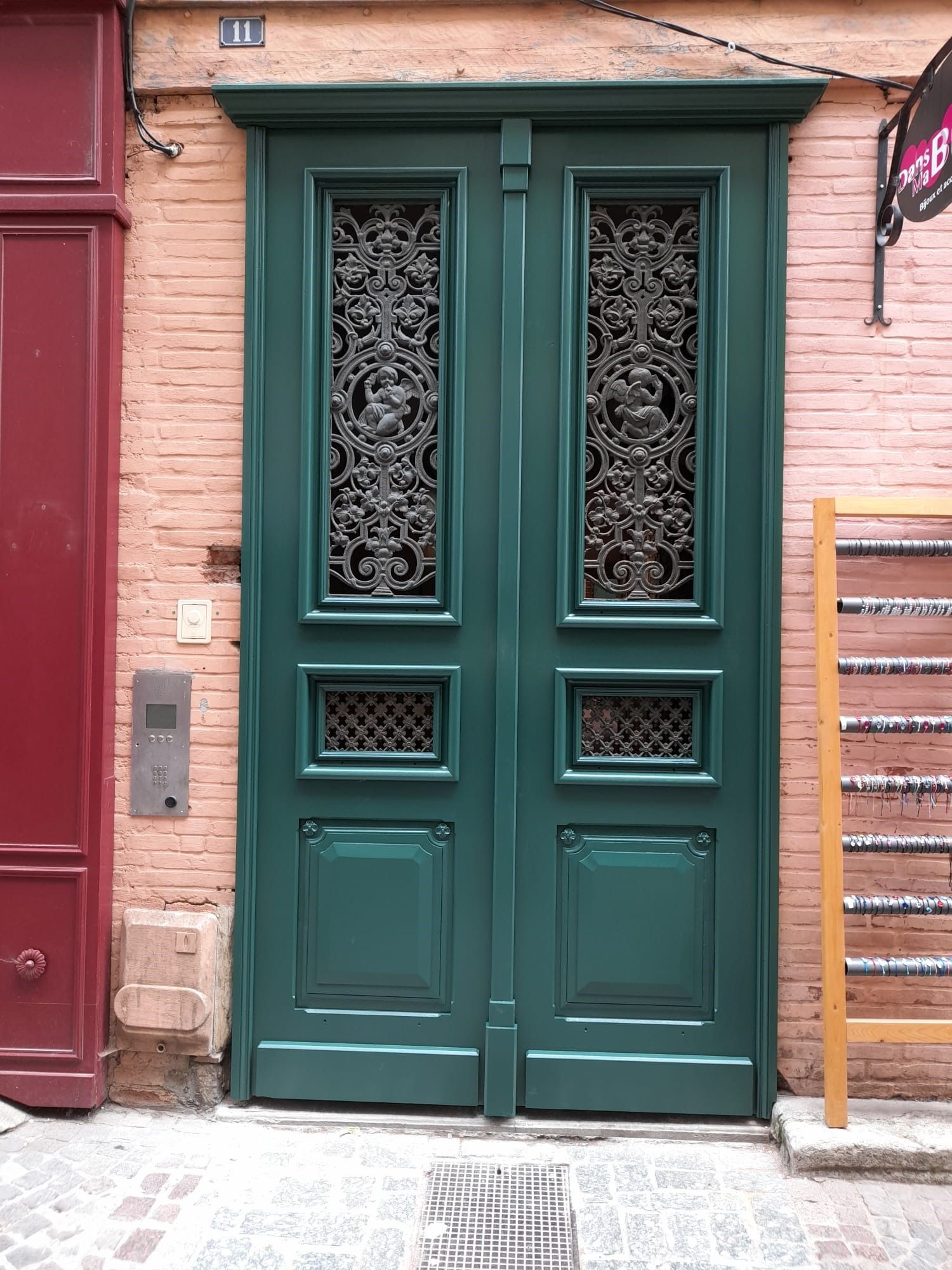 Porte double verte avec grilles métalliques ornées, placée dans un mur de briques entre une porte rouge et une vitrine en bois.
