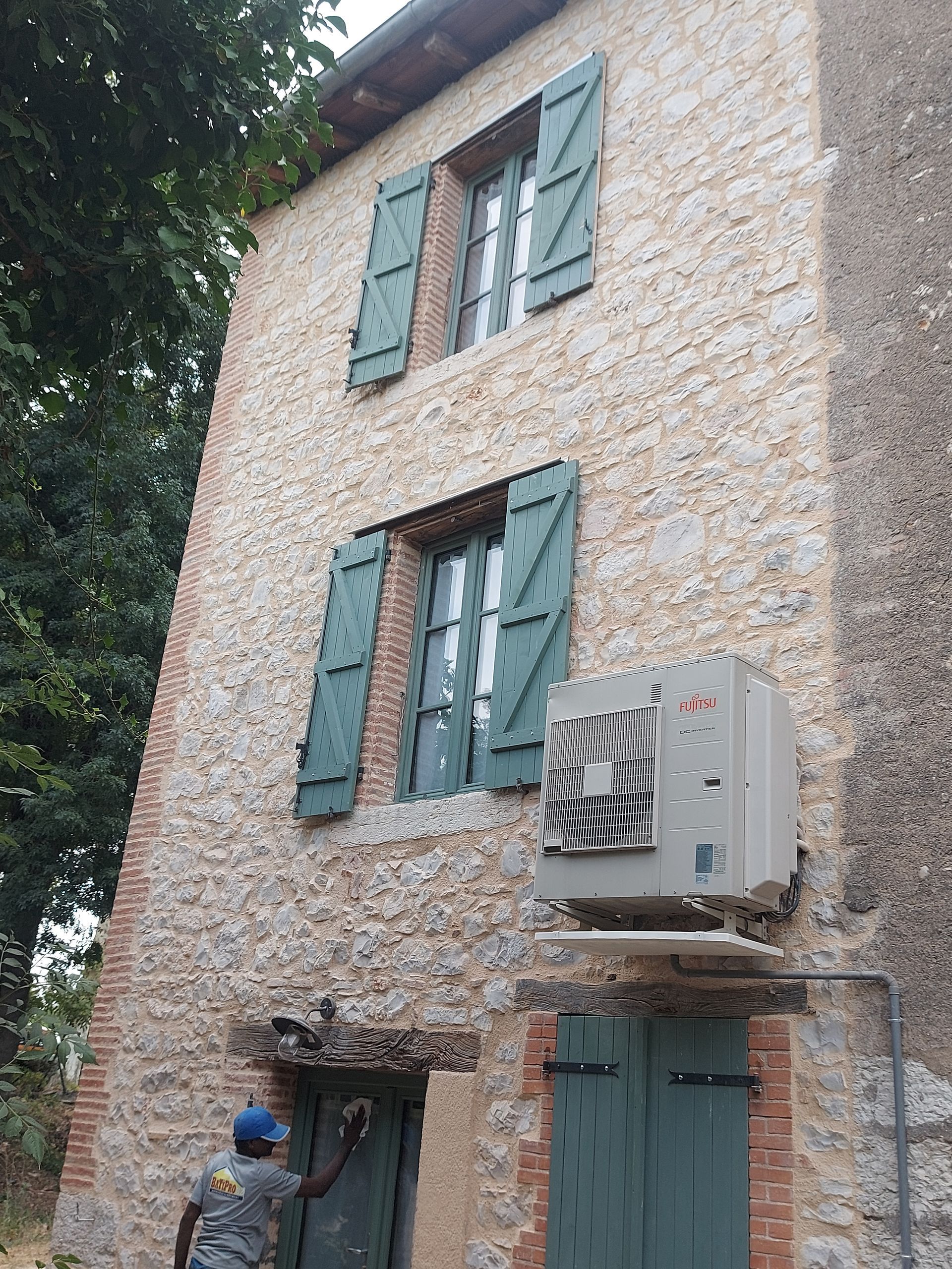 Bâtiment en pierre avec volets verts, unité de climatisation et une personne travaillant sur la porte.