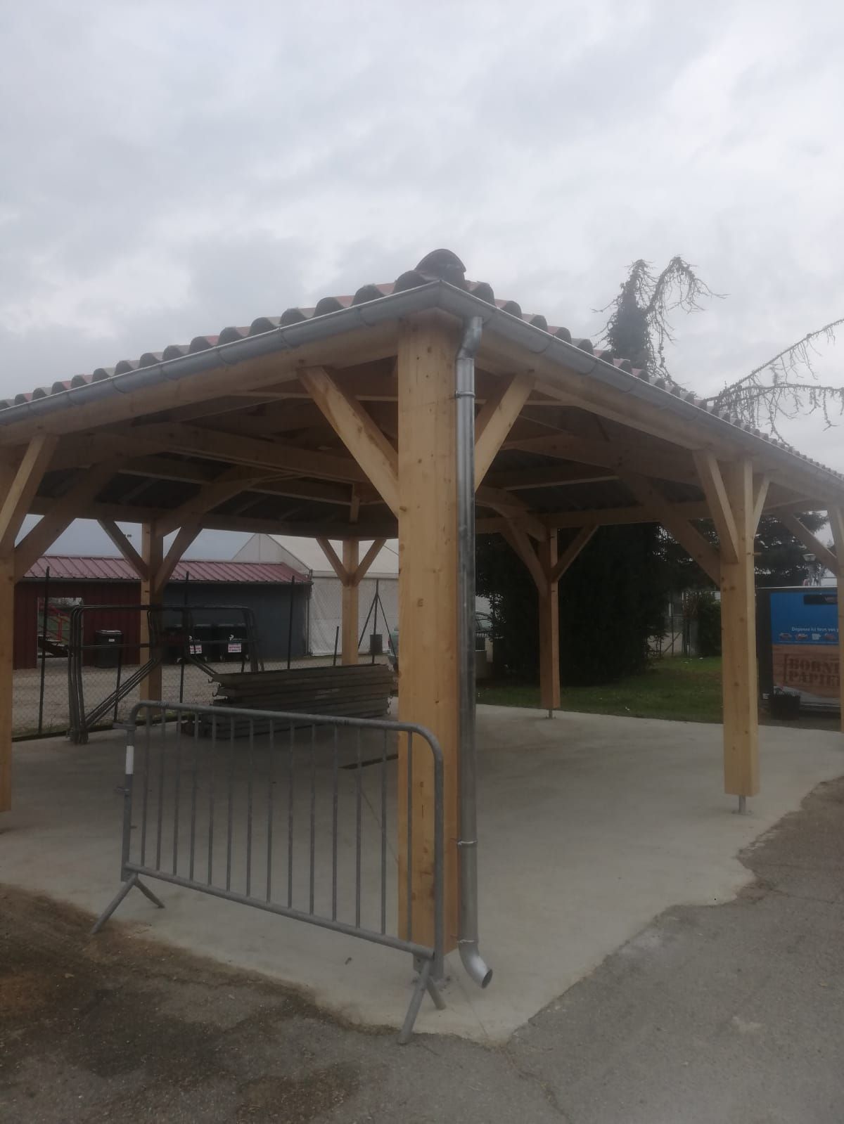 Pavillon en bois avec un sol en béton, une barrière en métal et un ciel gris couvert.