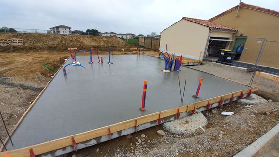 Chantier de construction avec murs en briques rouges, fondations en béton et ciel bleu. Une personne travaille dans une embrasure de porte.
