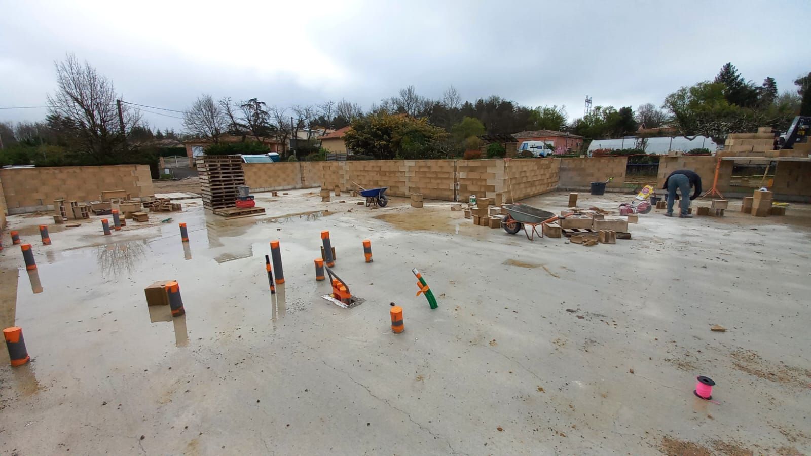 Chantier de construction avec murs de briques en construction sur une fondation en béton humide; plusieurs outils et ouvriers sont visibles.